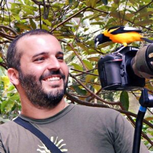 Leonardo Merçon, fotógrafo de natureza sorri enquanto um passaro chamado de "corrupião" ou "sofrê" pousa em sua câmera, interagindo com o fotógrafo.