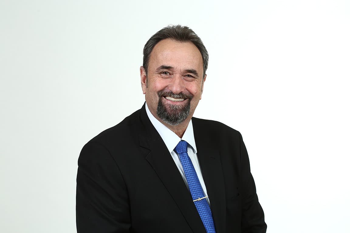 Foto de um homem branco, com cabelo preto, vestindo um terno preto e uma gravata azul